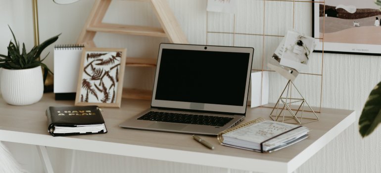 A clean and organized home office setup as part of New Year’s resolutions for home organization, featuring a laptop on a white desk with notebooks, a planner labeled '2024', and decorative elements such as a potted plant, a pinboard, and geometric desk accessories, all embodying a minimalist and clutter-free workspace.